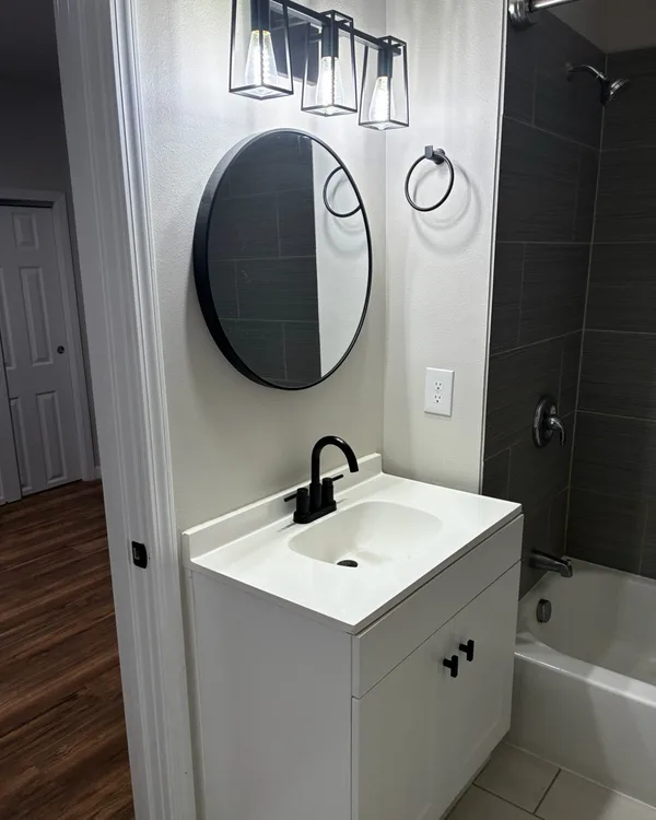 White bathroom vanity with a round mirror and black fixtures, beside a tiled tub and shower